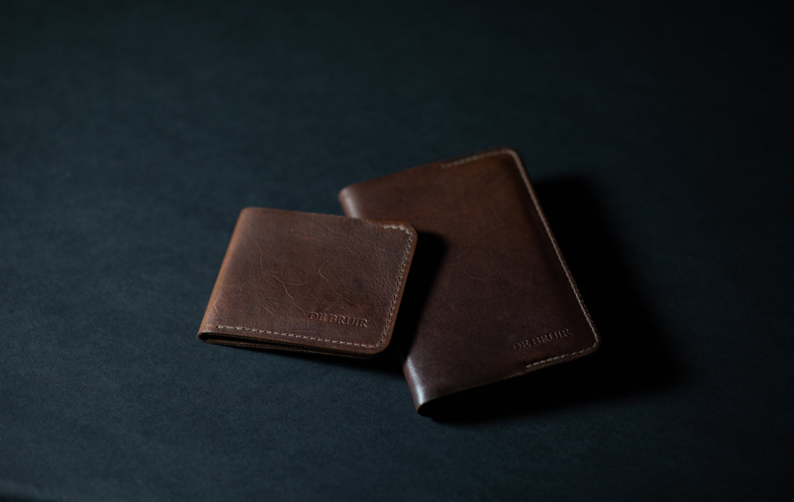 Two leather wallets, smaller on partly on top of larger, dark background